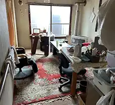 札幌市南区の1LDKアパートにて、引越し前に残された家具や家電、生活雑貨が室内に充満している状態。赤い絨毯の上にデスクや椅子、棚が置かれ、生活感が強く残っている作業前の様子。