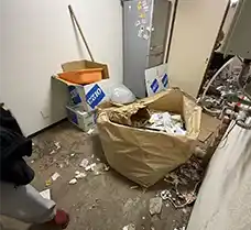 札幌市白石区のアパート4階、作業前のゴミ屋敷状態の室内。床一面に絨毯が見えないほどゴミや衣類が散乱し、デスクや棚の上にも生活用品が山積みになっている。窓際のストーブ周辺まで荷物で埋め尽くされた絶望的な様子。